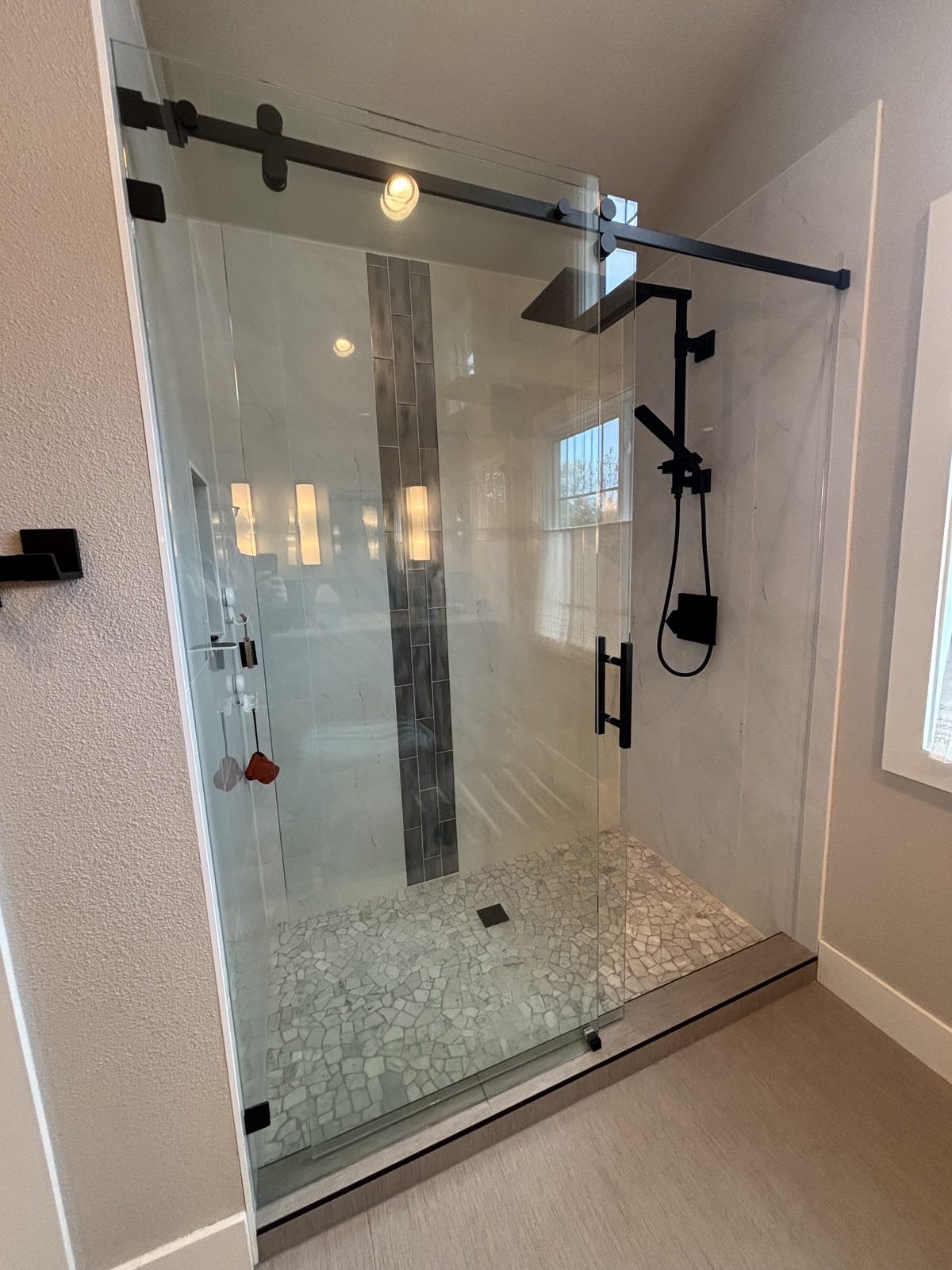 Finished walk-in shower with a frameless glass enclosure, matte black rainfall and handheld fixtures and a pebble tile floor